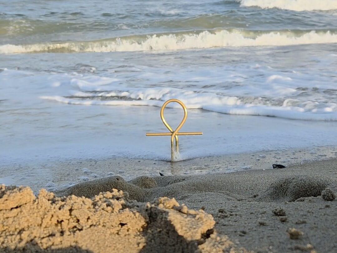 "Ankh met voeten in het zand en een terugtrekkende golf, symbool voor het loslaten van het negatieve, heling en herstel.